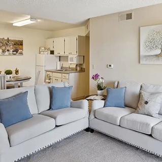 A cozy living room with two light gray sofas and blue cushions, a small side table with flowers, and an open kitchen with white cabinets and appliances in the background. A round dining table and wall art are also visible.