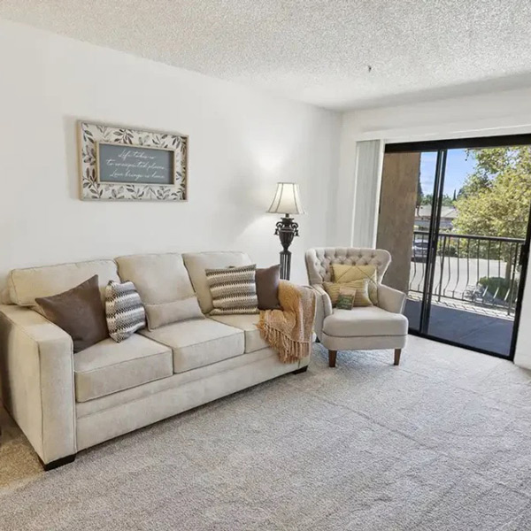 A bright living room with a beige sofa, accent pillows, a tufted armchair, a floor lamp, framed wall art, and a sliding glass door opening to a balcony with trees visible outside.
