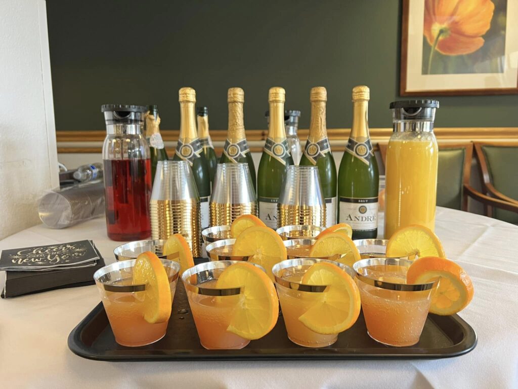 A tray of cocktails garnished with orange slices sits on a table alongside bottles of Andre champagne, clear cups, a carafe of red liquid, and a carafe of orange juice. A floral painting hangs on the wall behind.
