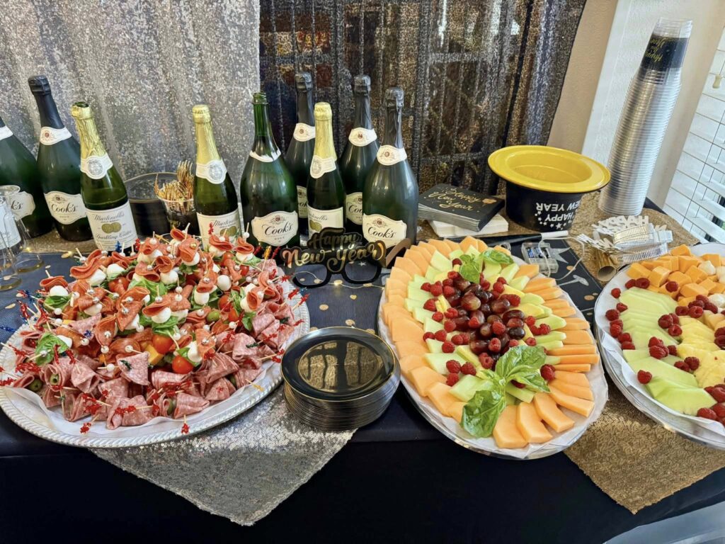 A festive table displays assorted meats on skewers, sliced cheeses, melon, and grapes, with bottles of sparkling wine and plastic cups behind. Gold and black New Year's Eve decorations add a celebratory touch.
