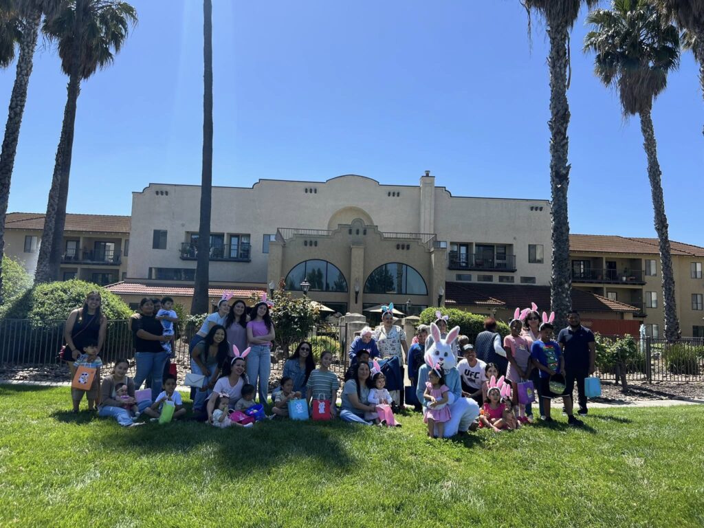 A large group of adults and children pose together on a sunny lawn in front of a tan building with palm trees. Many children hold colorful baskets and some people wear bunny ears.