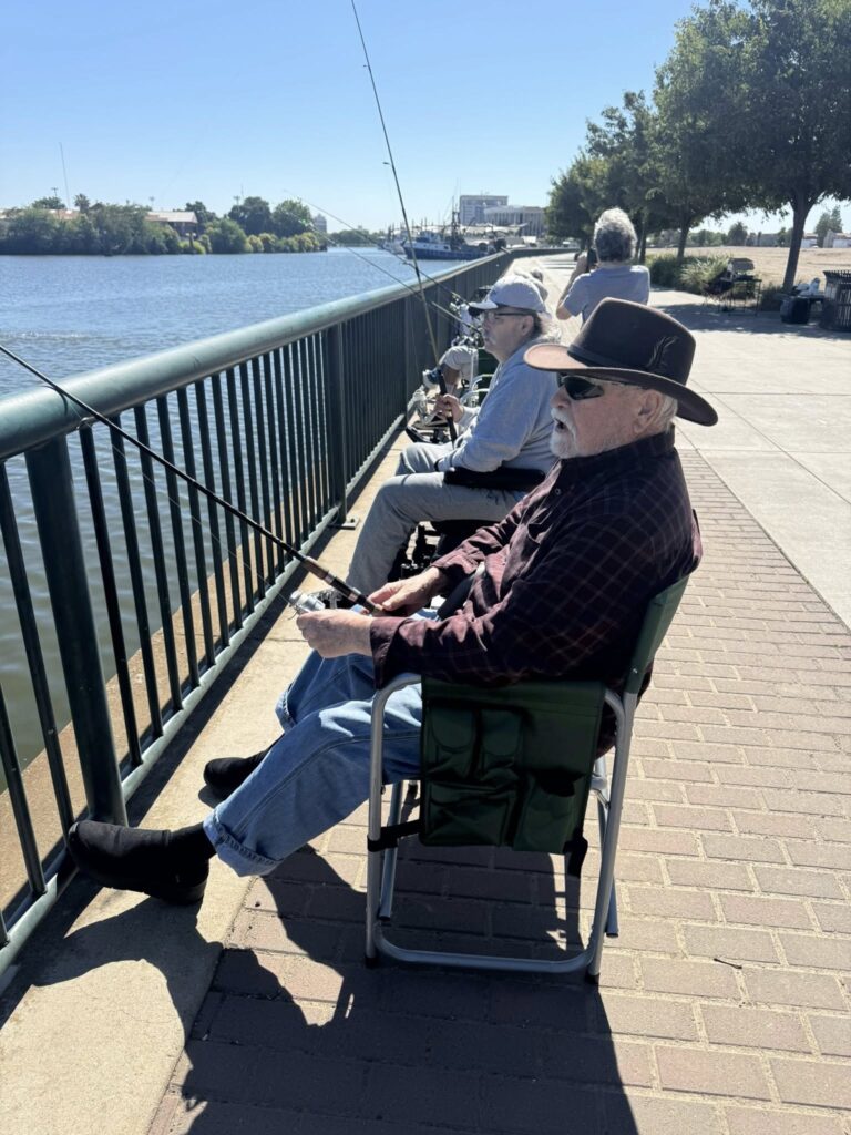 Three older men sit in chairs along a riverside promenade, fishing with rods over a green metal railing on a sunny day. Trees line the walkway, and water and boats are visible in the background.