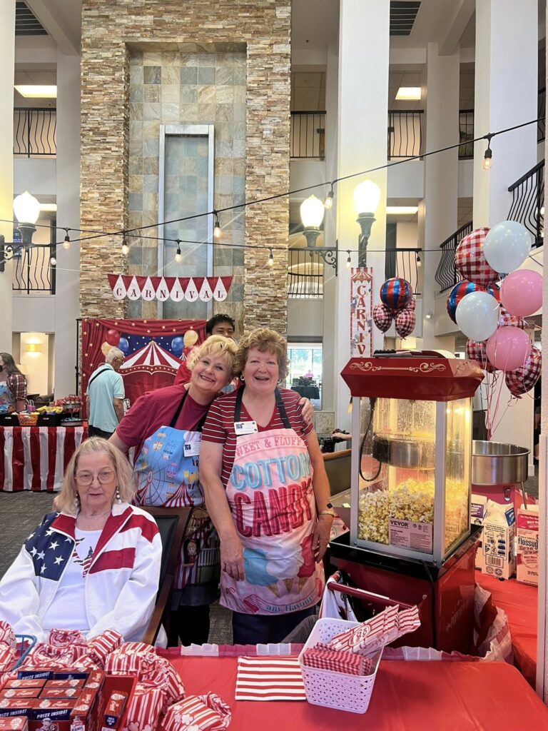 Three women at a festive indoor event, with one seated and two standing behind a popcorn and cotton candy stand. The setting is decorated with red, white, and blue balloons, banners, and tablecloths.