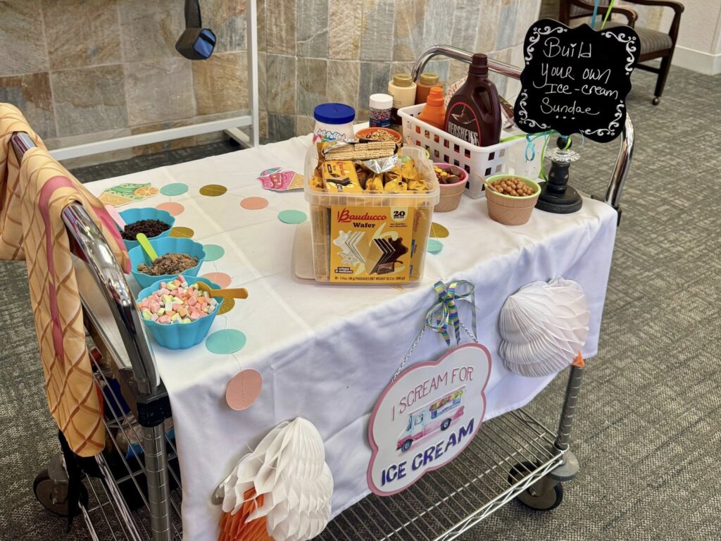 A cart decorated with ice cream-themed items holds various toppings, sauces, cones, bowls, and a sign reading “Build your own Ice Cream Sundae.” A cloth with “I Scream for Ice Cream” hangs on the front.