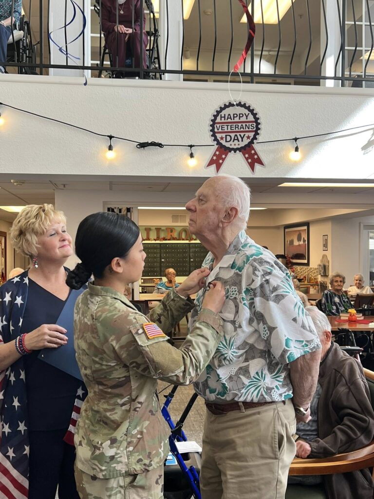 A woman in U.S. military uniform pins a medal on an elderly man’s shirt at a Veterans Day event, with onlookers and festive decorations in the background. A “Happy Veterans Day” sign hangs above them.