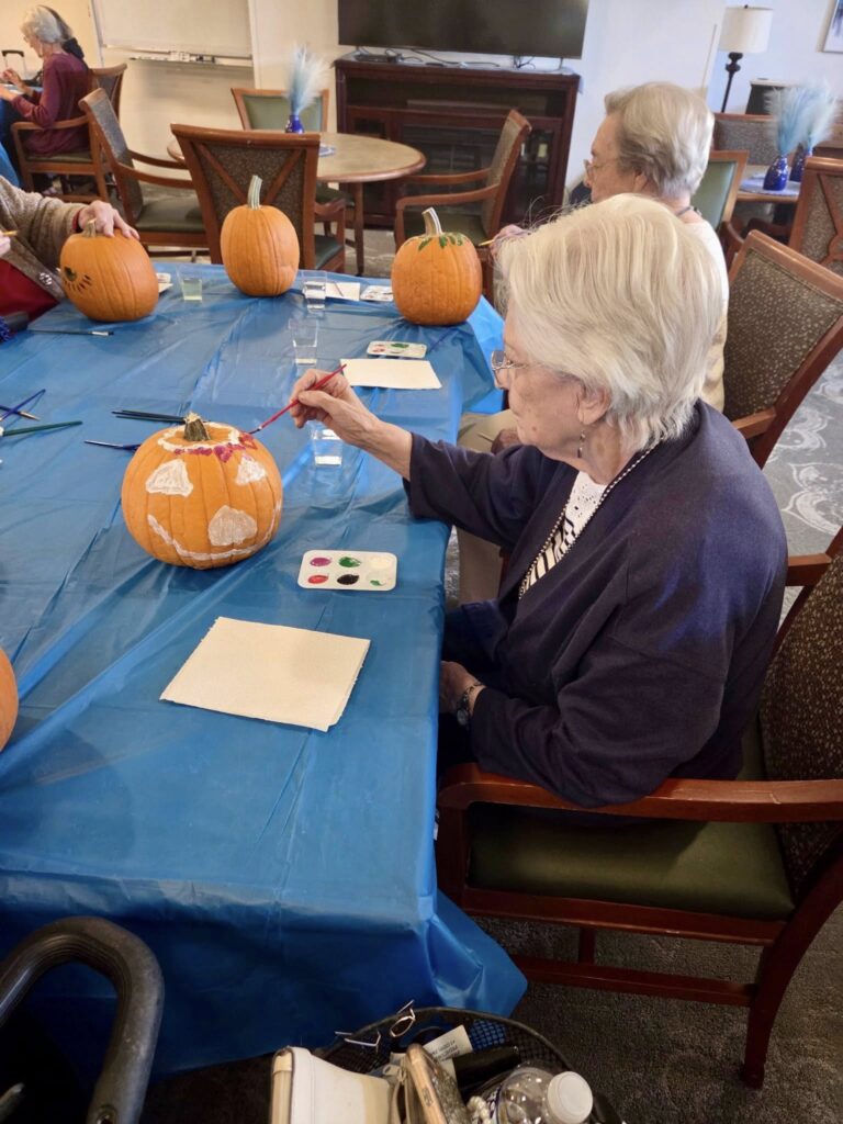 Two elderly women sit at a table covered with a blue tablecloth, painting pumpkins with brushes. Paint palettes and paper towels are on the table, and several unpainted pumpkins are nearby.