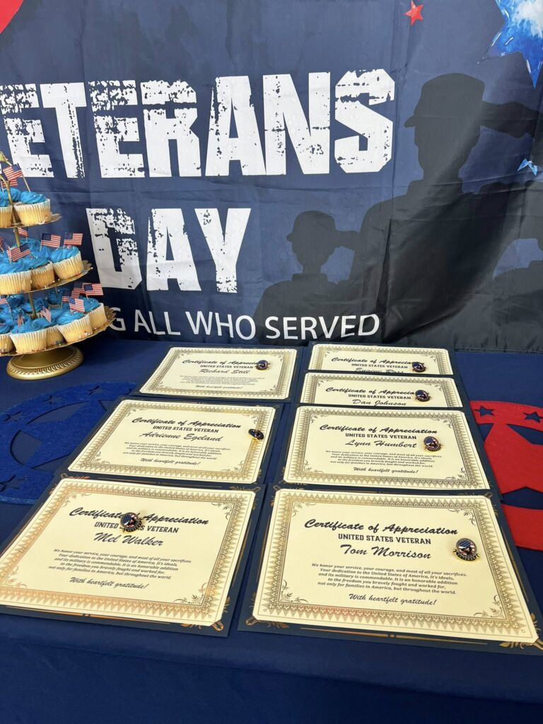 A table displays six "Certificate of Appreciation" awards for veterans on a blue tablecloth, with a stand of cupcakes and a "Veterans Day—Honoring All Who Served" banner in the background.