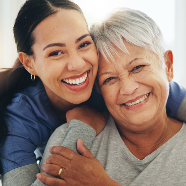 A young woman and an older woman are smiling and embracing closely, showing happiness and affection. The older woman has short gray hair, while the younger woman has long dark hair and wears a blue top.