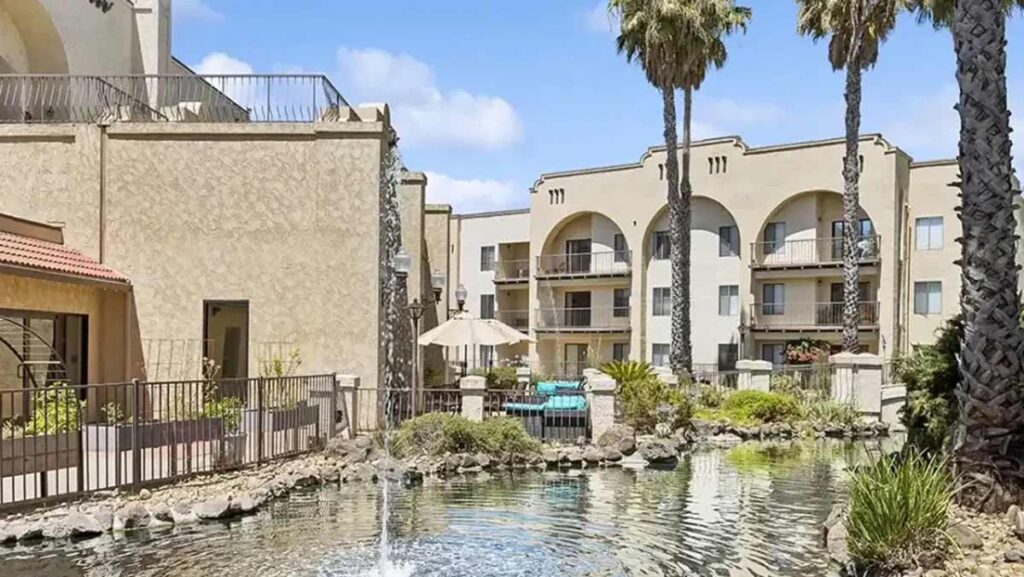 A courtyard with a small pond and fountain surrounded by palm trees and beige apartment buildings with balconies and arched windows, under a blue sky.