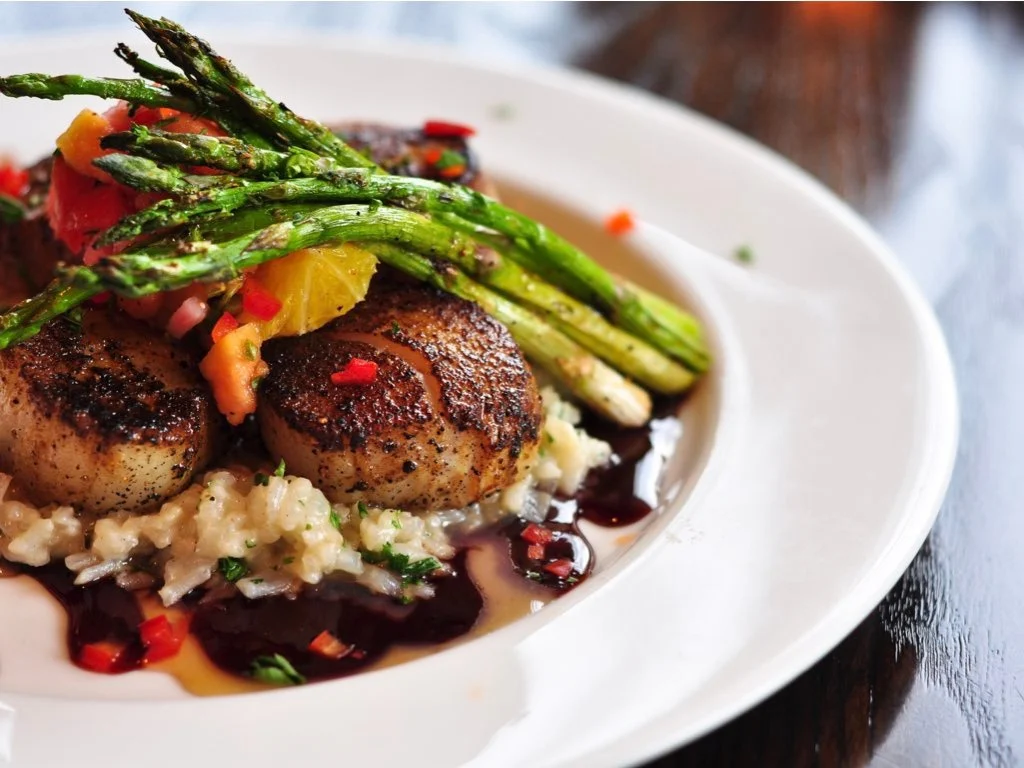 Seared scallops served on creamy risotto, topped with grilled asparagus, diced red peppers, and a citrus garnish, plated with two sauces on a white dish.