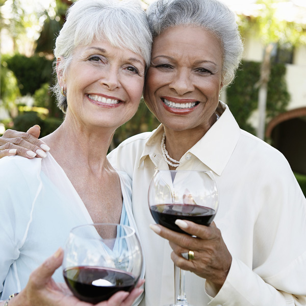Two older women smiling and standing close together outdoors, each holding a glass of red wine, with greenery and sunlight in the background.