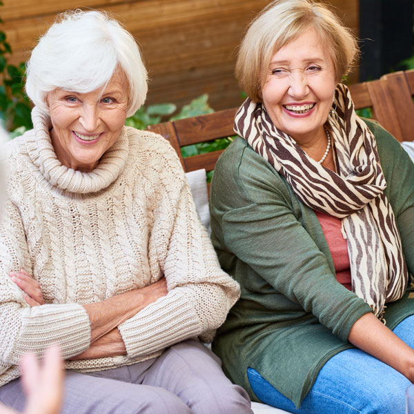 Two older women sitting on a bench outdoors, smiling and laughing together. One is wearing a cream sweater, the other a green top with a zebra-striped scarf. They appear to be enjoying a pleasant conversation.