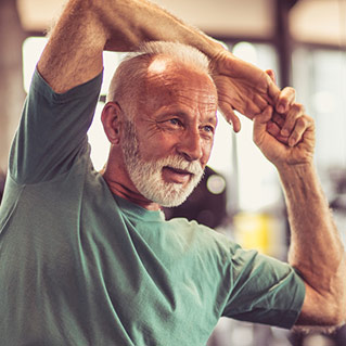 An older man with gray hair and a beard stretches his arm over his head while smiling. He is wearing a green t-shirt and appears to be in a gym or fitness center.