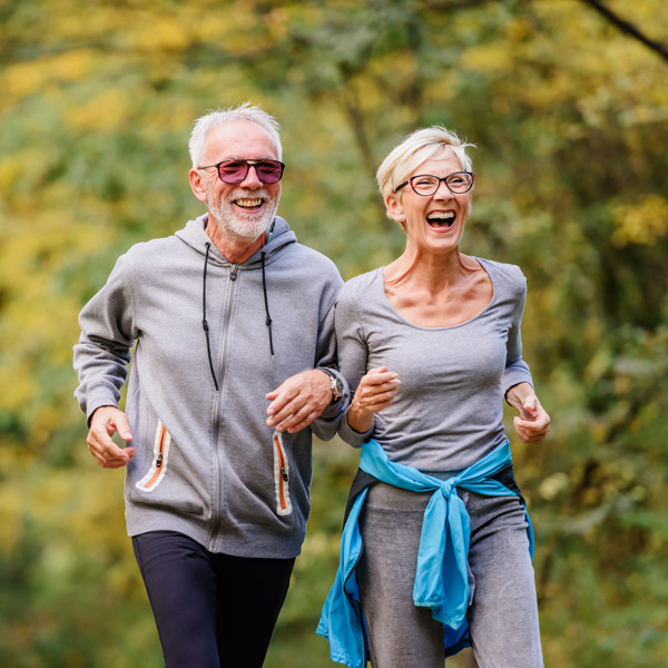 An older man and woman, both wearing glasses and gray activewear, are jogging outdoors together, smiling and looking happy. Trees with green and yellow leaves are in the background.
