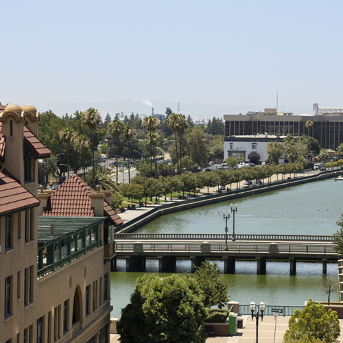 A cityscape featuring a tree-lined canal, a bridge crossing over it, and several buildings with red-tiled roofs and palm trees under a clear sky.