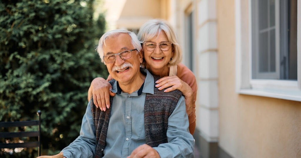 An older couple smiles warmly outdoors; the woman stands behind the seated man, resting her hands on his shoulders. Both wear glasses and casual clothes, with greenery and a building in the background.