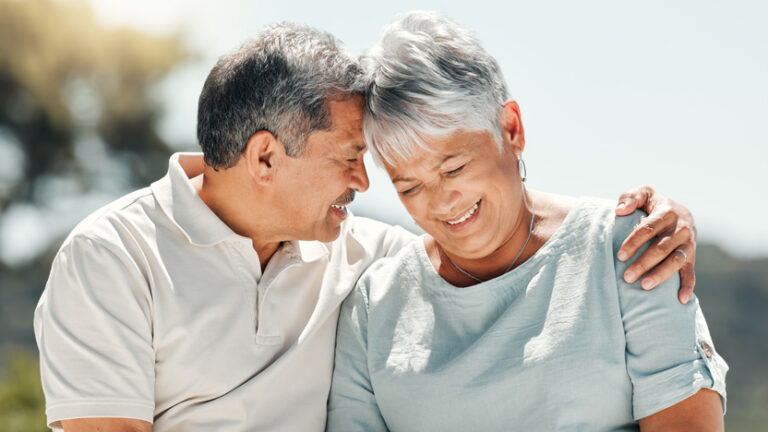 An older couple sits close together outdoors, smiling and embracing. The man is wearing a light polo shirt and the woman is in a light blouse. They look happy and affectionate, enjoying a warm moment—an inspiring scene for solo agers seeking meaningful connections in later life.